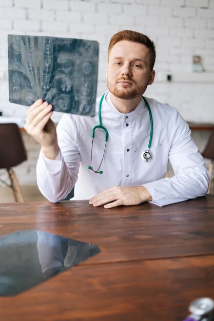 Disparo vertical de un médico masculino serio con uniforme blanco que examina la tomografía computarizada del cerebro sentado en el escritorio en la oficina del hospital