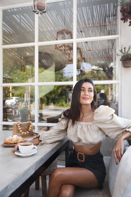 Disparo vertical de una mujer en traje de moda sentado zona de café con café y croissant, mirando a la calle.