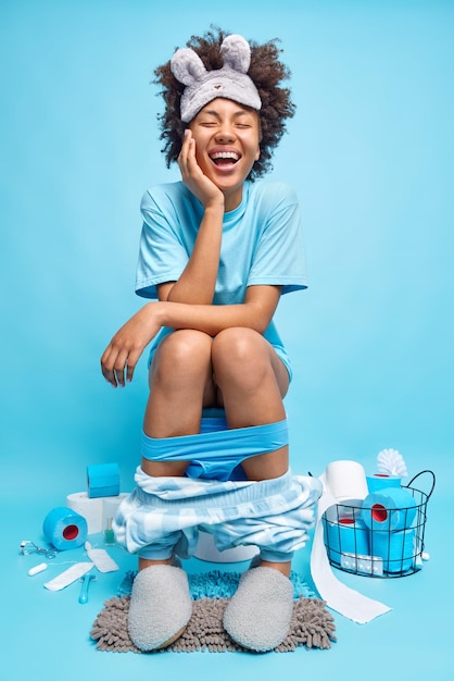 Disparo vertical de optimista mujer de pelo rizado vestida con pijama se ríe positivamente se revela a sí misma en la taza del inodoro aislado sobre fondo azul. Concepto de rutina diaria y necesidades humanas.