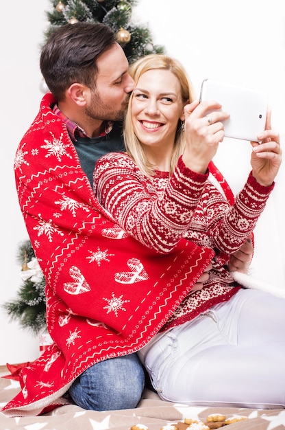 Disparo vertical de una pareja feliz con una manta roja tomando un selfie con un árbol de Navidad