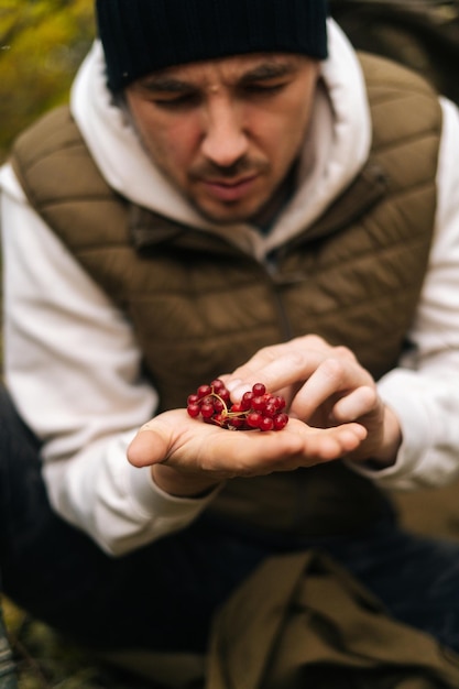 Disparo vertical de un superviviente del hambre pensativo sentado bajo un árbol en el bosque y sosteniendo bayas rojas silvestres en las manos mirándolas en un día lluvioso nublado