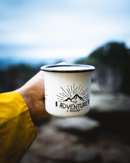 Disparo vertical de un viajero sosteniendo una taza frente a la vista borrosa al aire libre durante una aventura