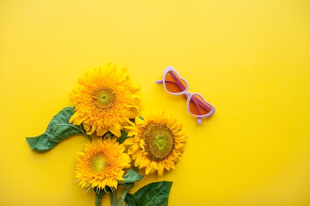 Foto disposición de gafas de sol rosas en forma de corazones y girasoles sobre un fondo amarillo brillante