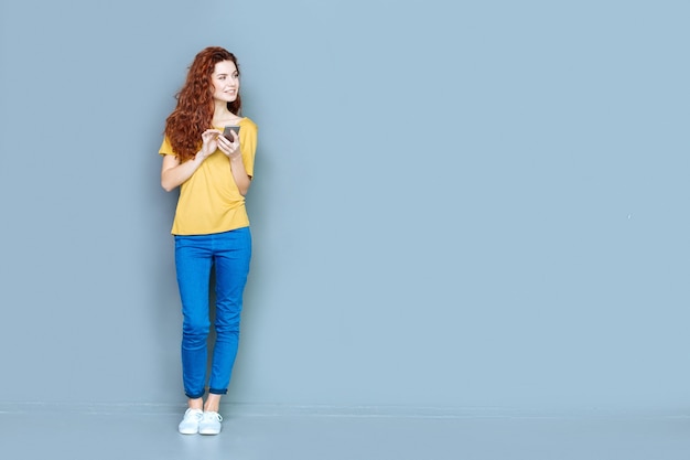 Dispositivo digital. mulher jovem e atraente e alegre em pé contra um fundo azul e sorrindo enquanto usa seu telefone celular