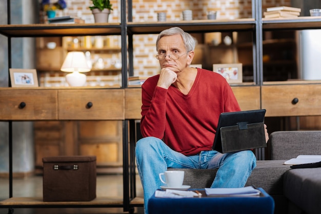 Dispositivo moderno. Agradable agradable hombre pensativo sosteniendo una mesa y mirándote mientras sostiene su barbilla