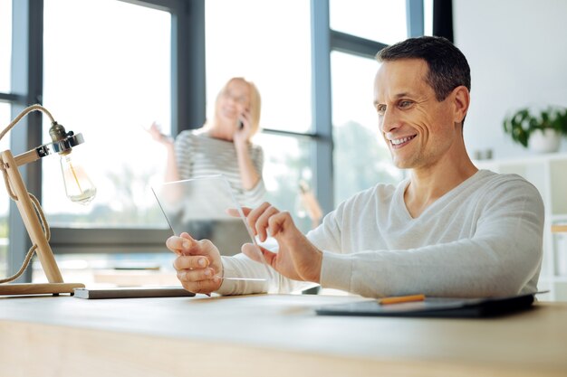 Dispositivo moderno. encantado, bonito e bonito homem segurando um tablet e usando-o enquanto está sentado à mesa em seu escritório