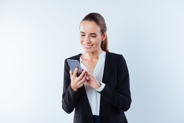 Dispositivo moderno. mujer alegre feliz positiva mirando la pantalla de su teléfono inteligente y sonriendo mientras está de pie contra el fondo azul