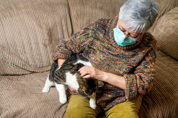 Distanciamiento social de coronavirus por brote de pandemia. Anciana con mascarilla protectora, permaneciendo en casa acariciando a su gato.