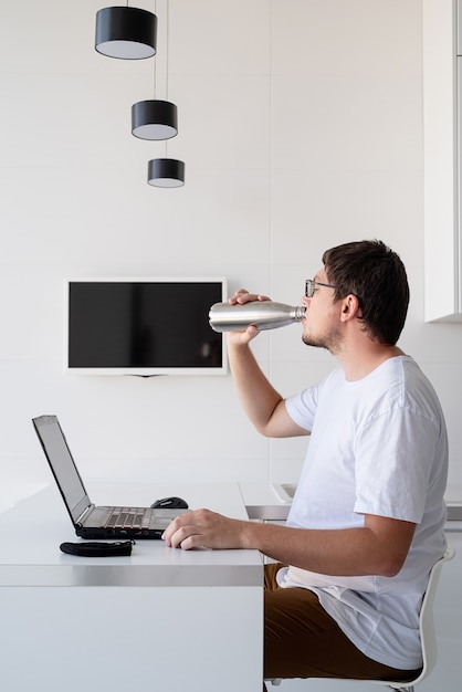 Distanciamiento social. E-learning y docencia. Hombre sonriente joven con camisa blanca usando laptop trabajando desde casa, bebiendo agua de termo