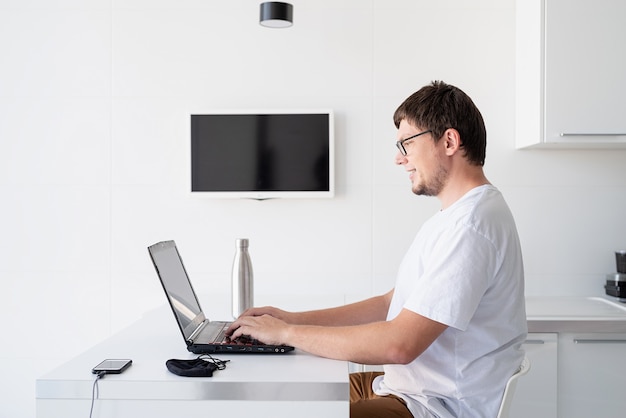 Distanciamiento social. E-learning y docencia. Hombre sonriente joven con camisa blanca usando laptop trabajando desde casa