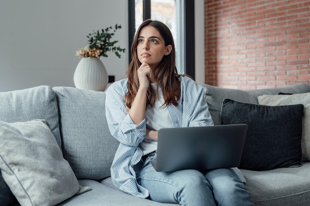 Distraída del trabajo mujer joven preocupada sentada en el sofá con la computadora portátil pensando en problemas mujer pensativa desmotivada mirando a la ventana sintiendo falta de energía haciendo tareas remotas independientes en homexA