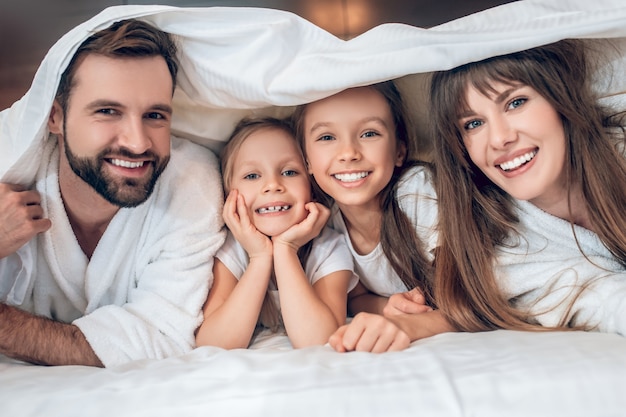 Diversão. Família sorridente com vestes brancas deitada na cama e se divertindo