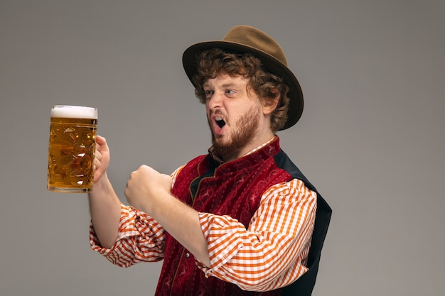Diversão. Homem feliz vestido com um traje tradicional austríaco ou bávaro, gesticulando com uma caneca de cerveja no fundo cinza do estúdio. Copyspace. A celebração, oktoberfest, festival, conceito de tradições.