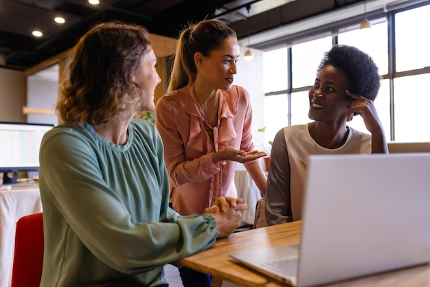 Foto diversas colegas femininas em discussão usando laptop em uma reunião casual de escritório.