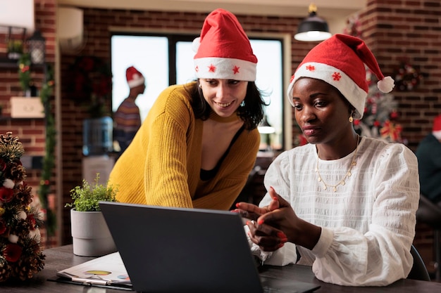 Diversas mulheres trabalhando em equipe no trabalho de inicialização com decorações festivas e árvore de natal, pessoas usando chapéu de papai noel no local de trabalho da empresa. Ornamentos de férias sazonais e luzes durante o inverno.