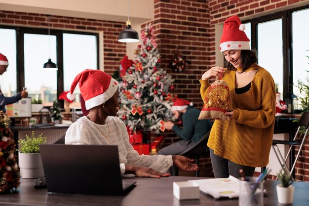 Diversas personas dando regalos en la oficina festiva para celebrar la nochebuena con decoraciones y luces de temporada. Las mujeres se sienten alegres después de intercambiar regalos en el espacio con árboles de Navidad y adornos.
