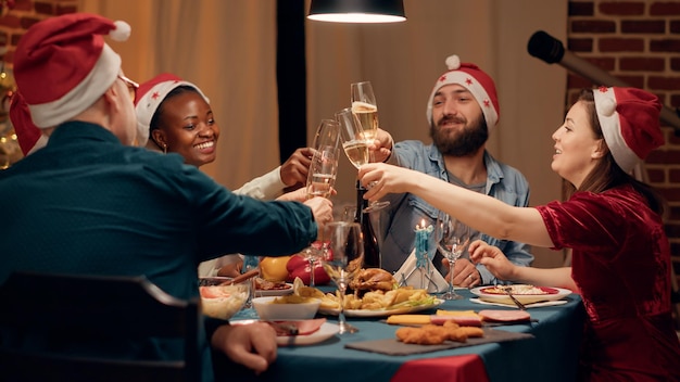 Diversas personas festivas que usan sombreros tradicionales mientras tintinean copas de champán en la cena de Navidad. Familia feliz celebrando las vacaciones de invierno con comida casera y vino espumoso. Disparo de mano