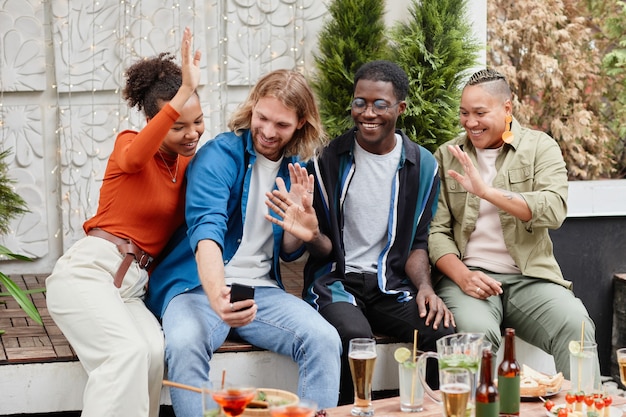 Diverse Gruppe junger Leute, die per Video-Chat in die Kamera winken, während sie sich bei einer Outdoor-Party auf dem Dach amüsieren