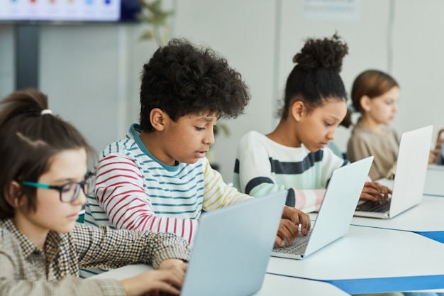 Diverse Gruppe kleiner Kinder, die Laptops benutzen, während sie im IT-Klassenzimmer in Reihe sitzen