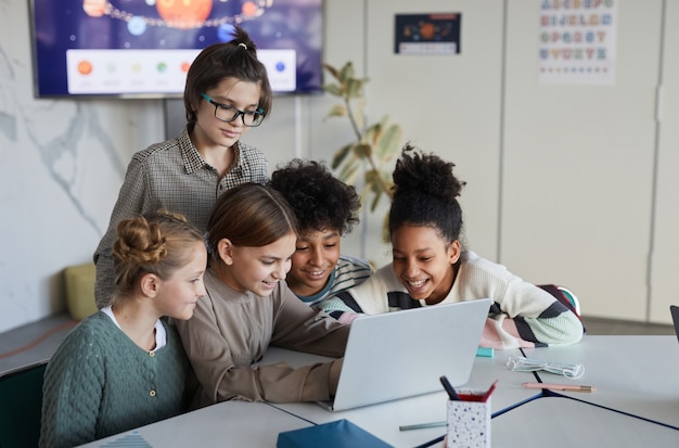 Diverse Gruppe von Kindern, die Laptop zusammen am Tisch in der modernen Schule benutzen, Platz kopieren