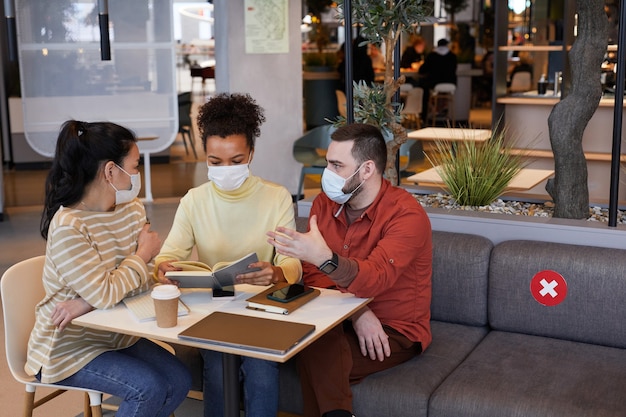 Diverse Gruppe von Menschen, die Masken tragen, während sie am Tisch im Café arbeiten, zusammen Geschäftstreffen mit Covid-Sicherheitsmaßnahmen, Platz kopieren