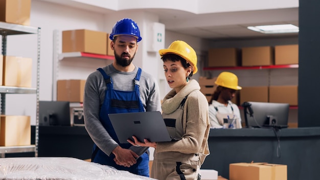 Foto diverse mitarbeiter im gespräch über lagerlogistik im depot, ware in kartons auf regalen im lagerraum platziert. mann und frau tragen overalls und hardhats mit laptop im lagerraum.