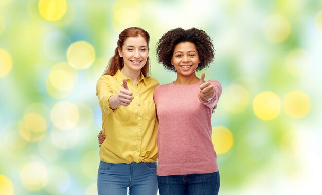 Foto diversidad, internacional, etnicidad, amistad y concepto de personas - mujeres sonrientes felices mostrando los pulgares hacia arriba y abrazándose sobre fondo de luces de vacaciones verdes