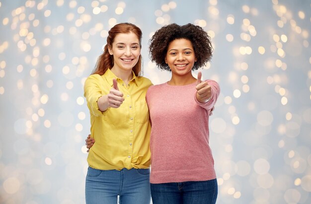 diversidad, internacionalidad, etnicidad, amistad y concepto de personas - mujeres felices y sonrientes mostrando los pulgares hacia arriba y abrazándose sobre las luces de las fiestas de fondo