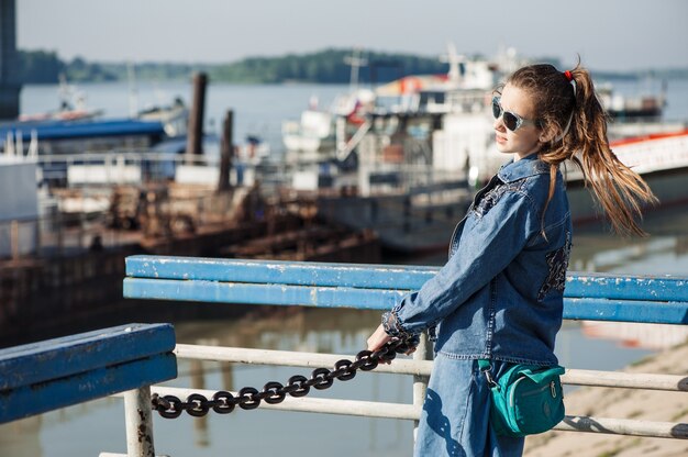 Diversión adolescente en vestido de mezclilla azul está caminando en la estación del río. Niño alegre haciendo pipí en el puerto fluvial. Chica con gafas de sol sujeta la cadena.