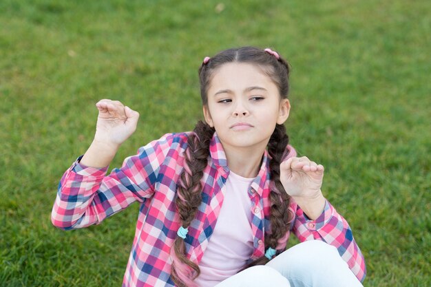 Diversión al aire libre de la pequeña niña relajarse en la hierba verde. Parques y exteriores. Naturaleza primaveral. Picnic de verano. Niña de la escuela pequeña con el pelo de moda divirtiéndose. infancia feliz. Niño gracioso con cara divertida. solo diversión.