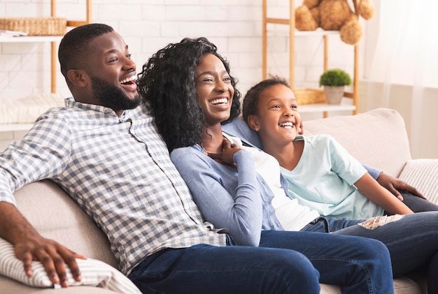 Diversión familiar. Los jóvenes padres afroamericanos y su pequeña hija se ríen mientras ven un programa de comedia en la televisión