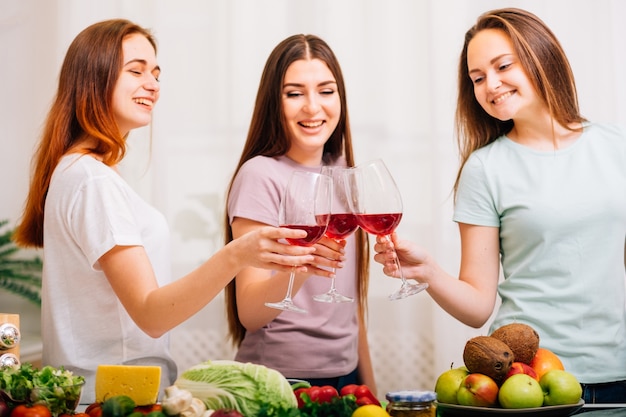 Diversión de fiesta femenina. Surtido de alimentos saludables. Mujeres jóvenes felices tintineando copas de vino tinto.