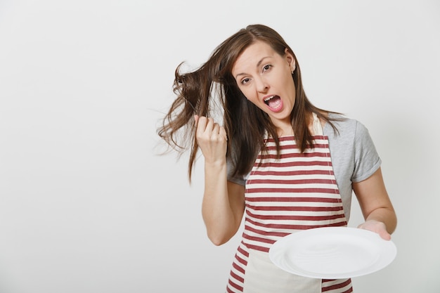 Diversión joven ama de casa morena loca en delantal de rayas camiseta gris aislado Ama de llaves mujer sosteniendo blanco plato vacío tenedor en el pelo como peine cepillo