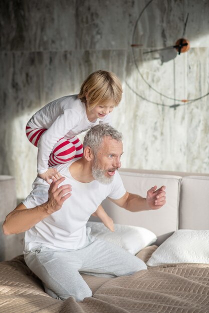 Diversión con papá. Niño feliz subiéndose a la parte de atrás del padre alegre canoso arrodillado en la cama