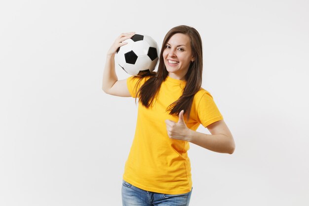 Diversión sonriente joven europea, aficionado al fútbol en uniforme amarillo sosteniendo un balón de fútbol mostrando los pulgares para arriba gesto aislado sobre fondo blanco. Deporte, jugar al fútbol, animar, concepto de estilo de vida de las personas de los fanáticos.