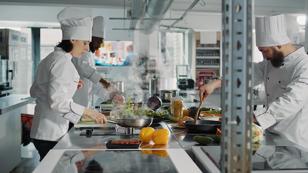 Diverso equipo de chefs preparando ingredientes de platos gourmet, cocinando deliciosa comida en la estufa. Personas con uniforme blanco que usan recetas profesionales para hacer comida de gastronomía orgánica con verduras.