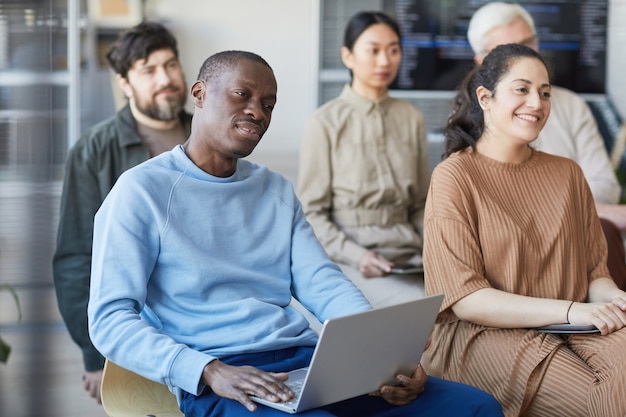 Diverso grupo de empresarios sentados en sillas en la audiencia y escuchando en una reunión o seminario, se centran en el hombre afroamericano con una computadora portátil en primer plano