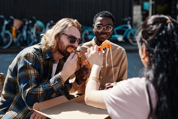 Diverso grupo de jóvenes disfrutando de la pizza al aire libre, escena iluminada por la luz del sol, espacio de copia