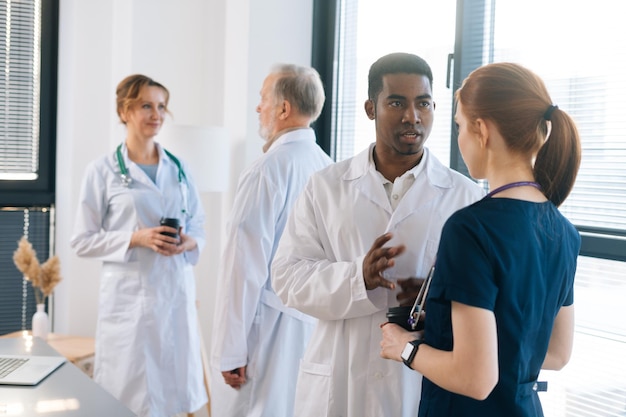 Diverso grupo de médicos profesionales masculinos y femeninos multiétnicos que tienen una conversación en la clínica de pie en el fondo de la ventana