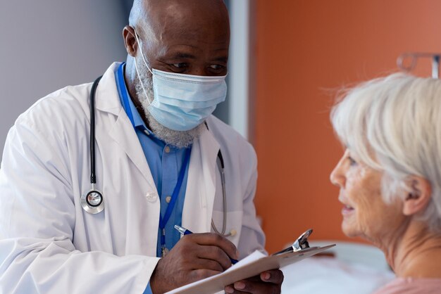 Diverso médico senior con mascarilla hablando con una paciente senior, tomando notas, copiando espacio. Servicios hospitalarios, médicos y sanitarios.