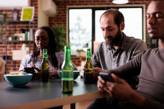 Diversos amigos sentados juntos en la mesa en la sala de estar mientras navegan por las redes sociales en los teléfonos inteligentes. Personas multiétnicas sentadas en casa mientras navegan por Internet en modernos dispositivos de pantalla táctil.