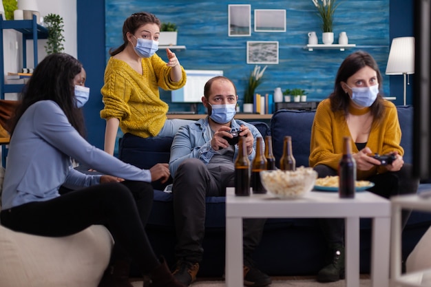 Diversos amigos tentando vencer jogando videogame usando o joystick se divertem usando uma máscara facial para evitar a propagação do coronavírus em tempos de surto global. Competição de jogos, cerveja e pipoca.