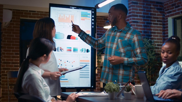 Diversos analistas que presentan visualización de estadísticas de marketing de la empresa en reuniones de negocios, empleados que escuchan la presentación. Trabajadores de oficina en la sala de juntas del espacio de coworking por la noche