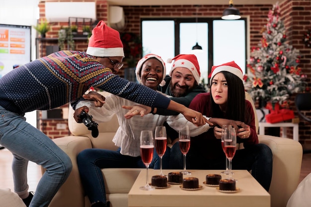 Diversos compañeros de trabajo jugando videojuegos en un evento de navidad, celebrando la festividad de vacaciones de invierno con colegas en el lugar de trabajo decorados con adornos de temporada. Divertirse jugando en la fiesta.