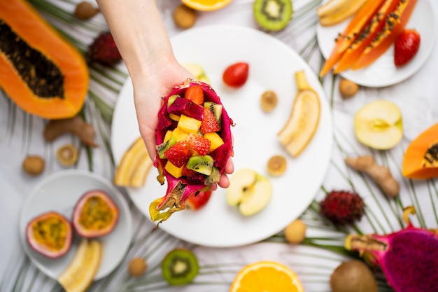 Diversos frutos maduros suculentos tropicais frescos no verão na mesa. Perto da mão feminina, segurando a metade da fruta do dragão cheia de salada de frutas. Postura plana, vista superior, foco seletivo. Lanche delicioso.