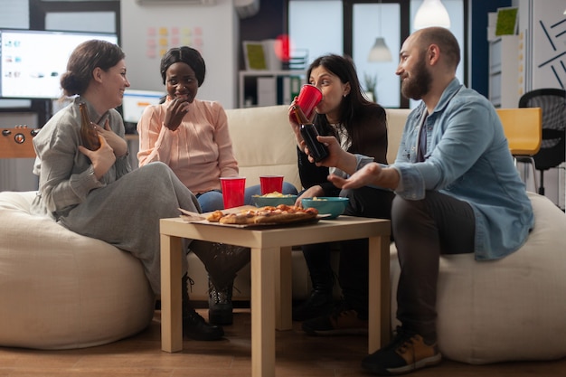 Diversos grupos de colegas desfrutam de bebidas de festa após o trabalho no escritório. Multi étnicos amigos alegres, sentados no sofá com salgadinhos, chips de pizza e copos de garrafas de álcool de cerveja na mesa.