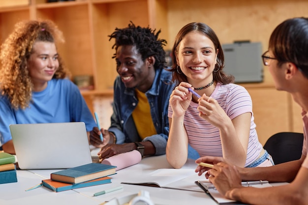 Diversos grupos de estudantes trabalhando juntos à mesa na biblioteca se concentram em uma jovem sorridente falando