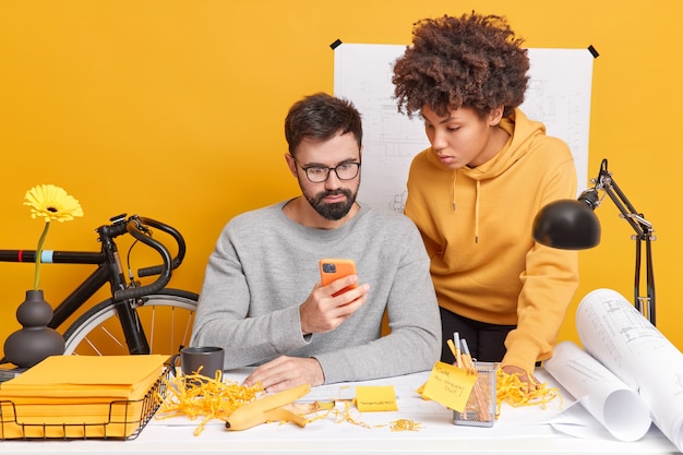 Diversos hombres y mujeres ocupados trabajan juntos en la oficina concentrados en el teléfono inteligente, aprenden información del sitio web, tienen expresiones atentas, comprueban el correo electrónico, desarrollan una nueva estrategia para el proyecto de ingeniería