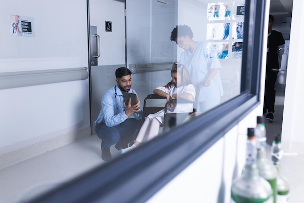 Foto diversos médicos masculinos y femeninos hablando con una niña en silla de ruedas usando una tableta en el pasillo del hospital