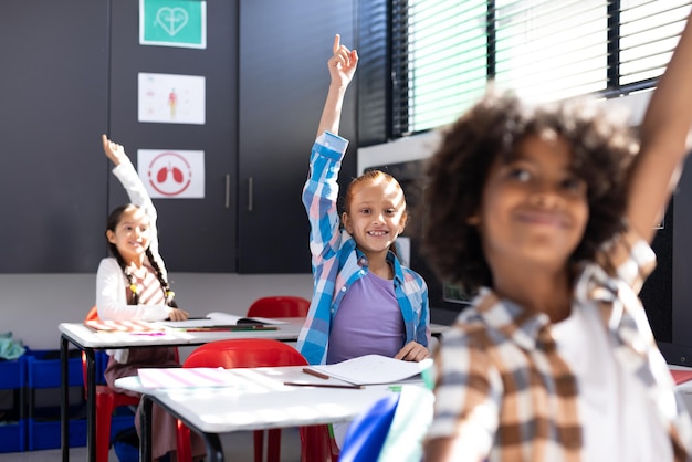 Diversos niños felices sentados en escritorios levantando las manos en el espacio de copia del aula de la escuela primaria